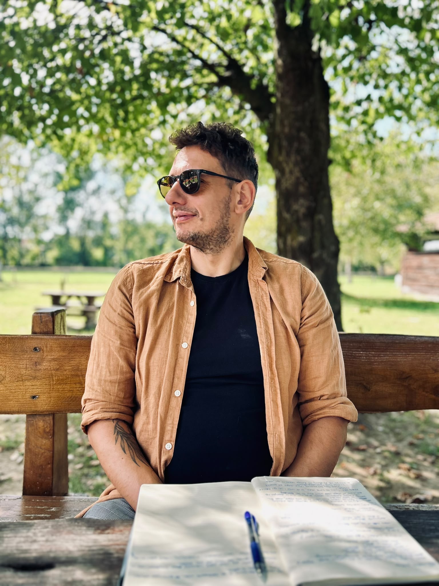 Filip Sardi on a wooden bench under a tree in sunglasses, an open notebook and pen on the table in front of him