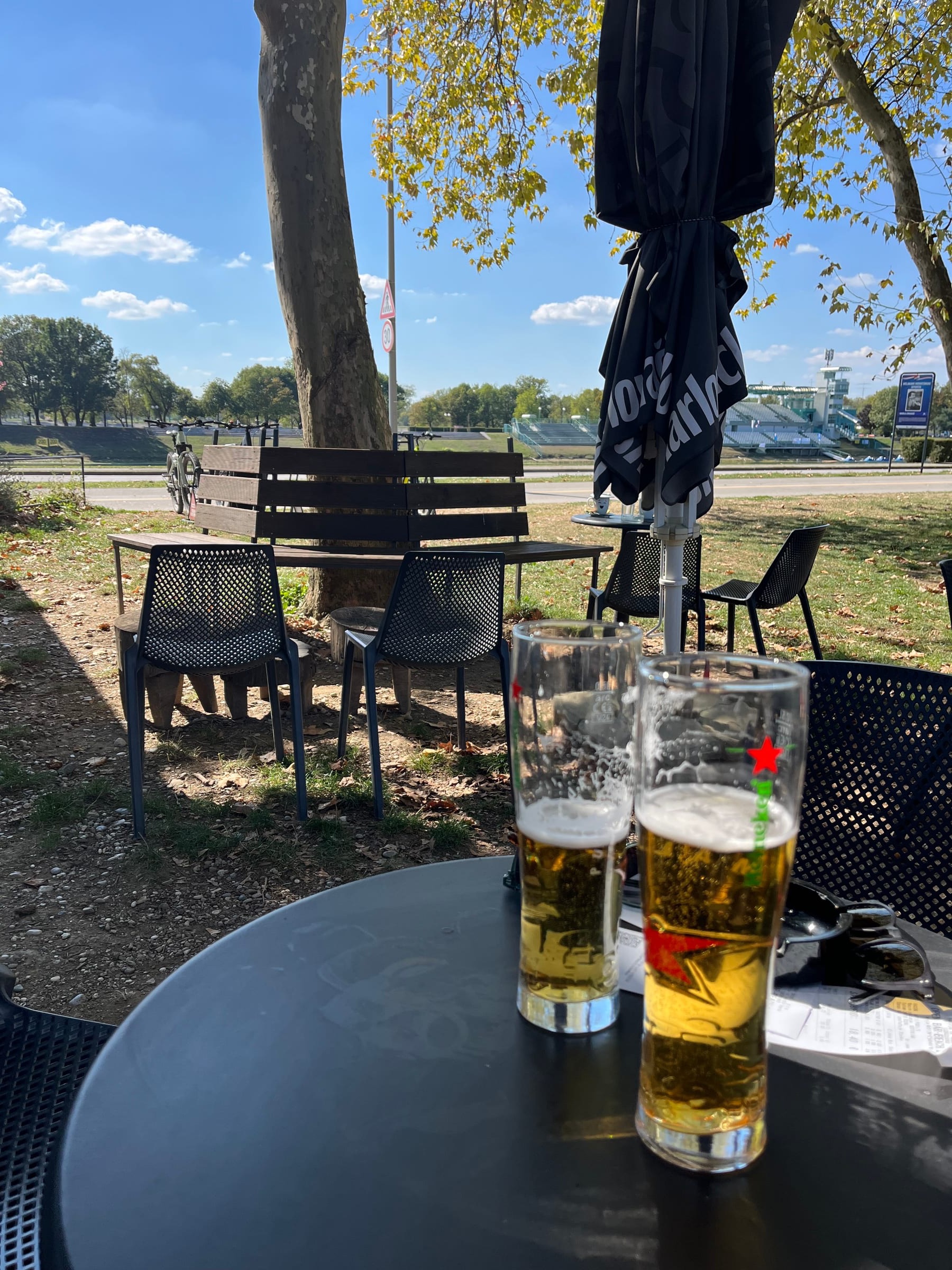 Two glasses of beer on a round black table at an outdoor bar under plane trees, the stadium stands in the distance