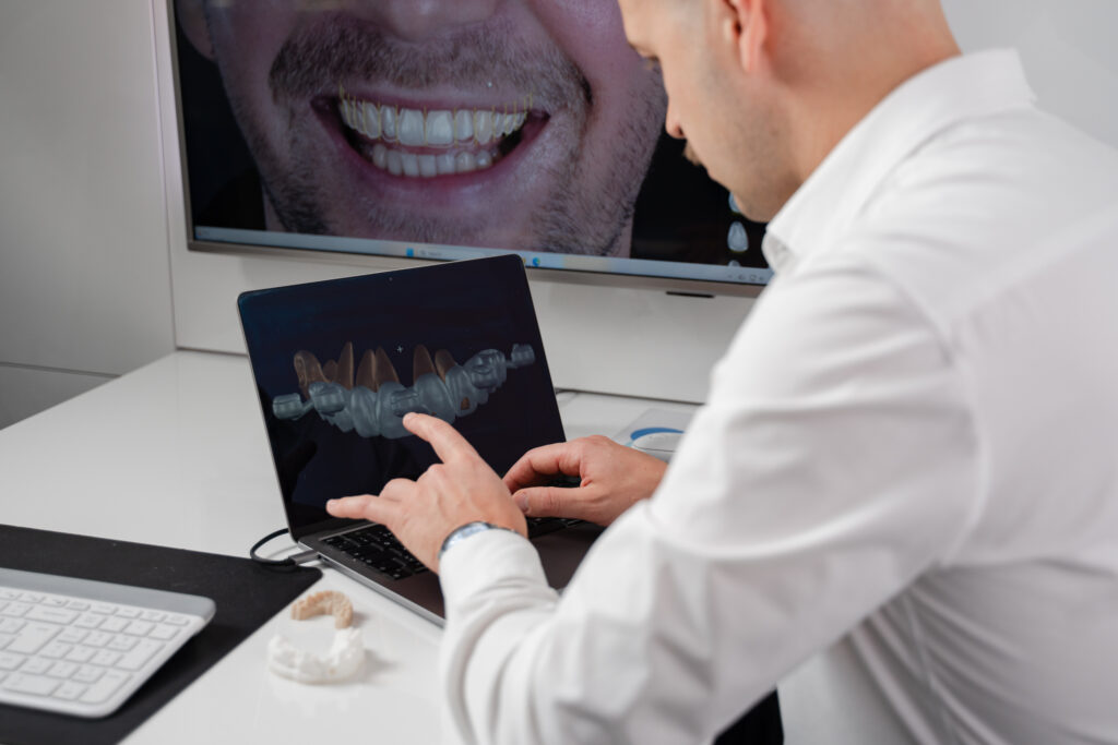 A dentist at a laptop pointing at a 3D scan of teeth, a patient's smile on the screen behind him